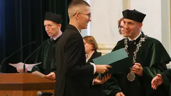 Na zdjęciu widzimy uroczystość wręczenia dyplomów. Młody mężczyzna, ubrany w elegancki garnitur, odbiera dokument z rąk Dziekana. Otrzymujący dyplom uśmiecha się, a w tle widać innych uczestników ceremonii, również ubranych w stroje akademickie.