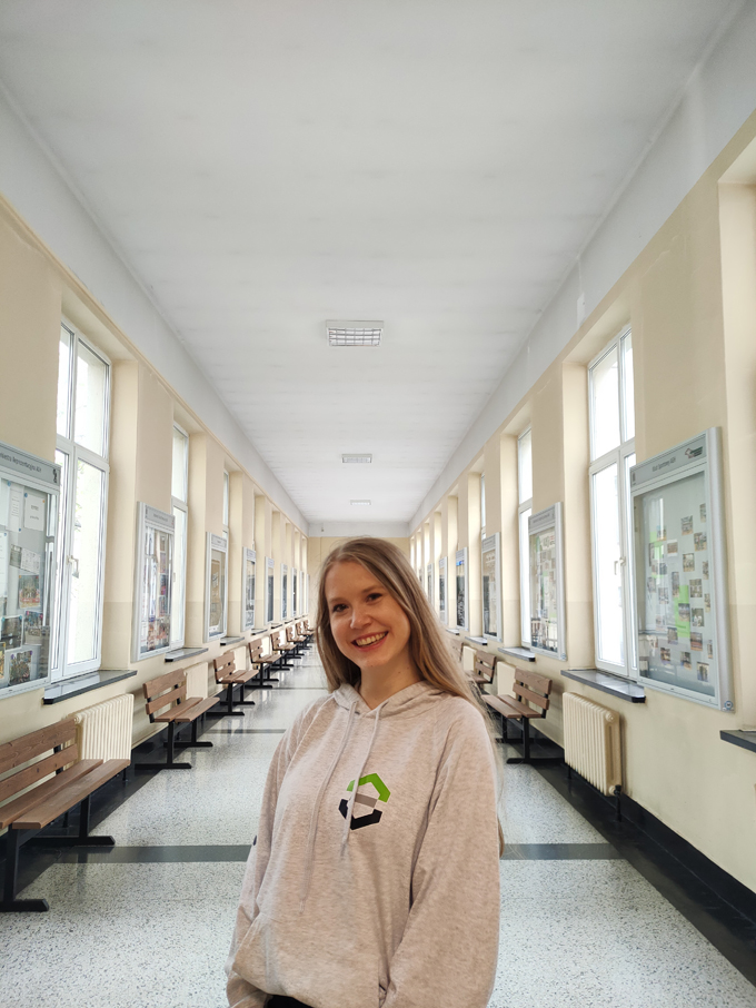 Usmiechnięta studentka w szrej bluzie wydziałowej stoi na środku korytarzu.