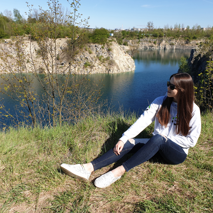 Studentka czarnych okularach przeciwsłonecznych siedzi w szarej bluzie wydziałowej na terenie zalewu Zakrzówek. W tle zallew Zakrzówek oraz wapienne klify.