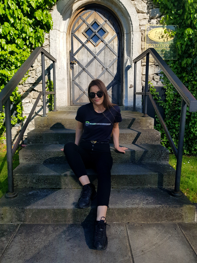 Studentka w czarnych okularach przeciwsłonecznych i czarnej bluzie wydziałowej siedzi na kamiennych schodach przed drewnianymi drzwiami porosniętymi po obu stronach winobluszczem.