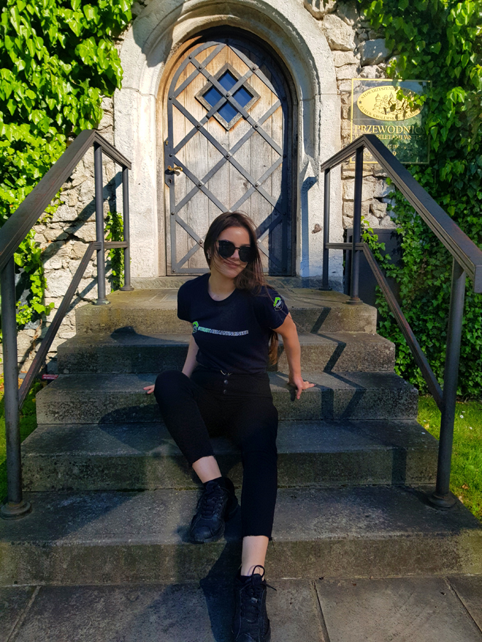 Studentka w czarnych okularach przeciwsłonecznych i czarnej bluzie wydziałowej siedzi na kamiennych schodach przed drewnianymi drzwiami porosniętymi po obu stronach winoroślą.