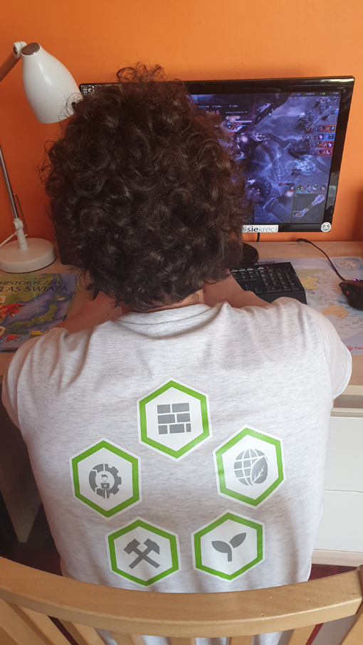 Mężczyzna w kręconych włosach ubramy w szary t-shirt gra na kompuerze. Na plecach bluzki znajduje się pięć zielonych heksów z symbolami kierunków kształćenia.
