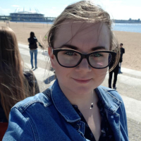 Uśmiechnięta młoda kobieta w okularach. W tle plaża, morze i spacerujący ludzie. Zdjęcie profilowe.