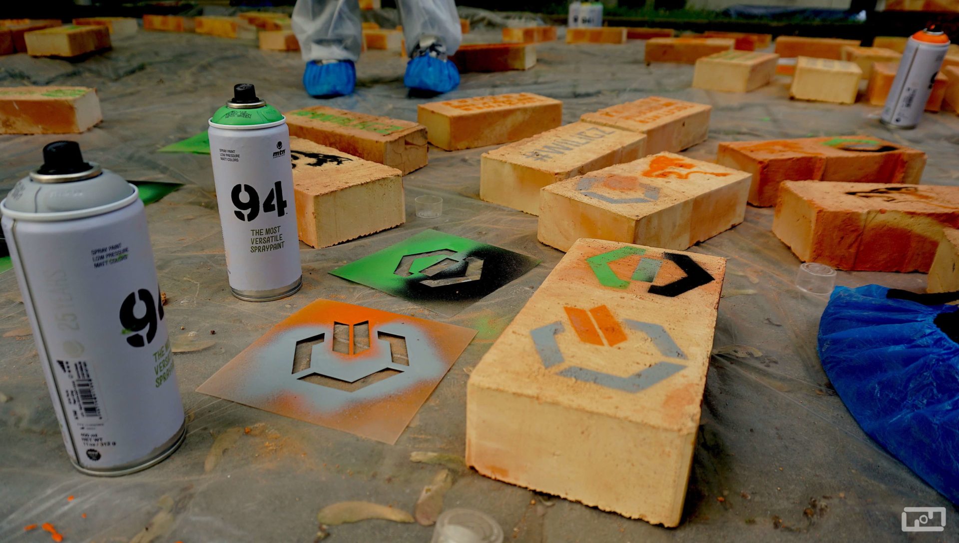 Pomalowane cegły leżące obok sprayów i szblonów przedstawiających logo WILiGZ raz logo WRSS leżą na folii malarskiej.