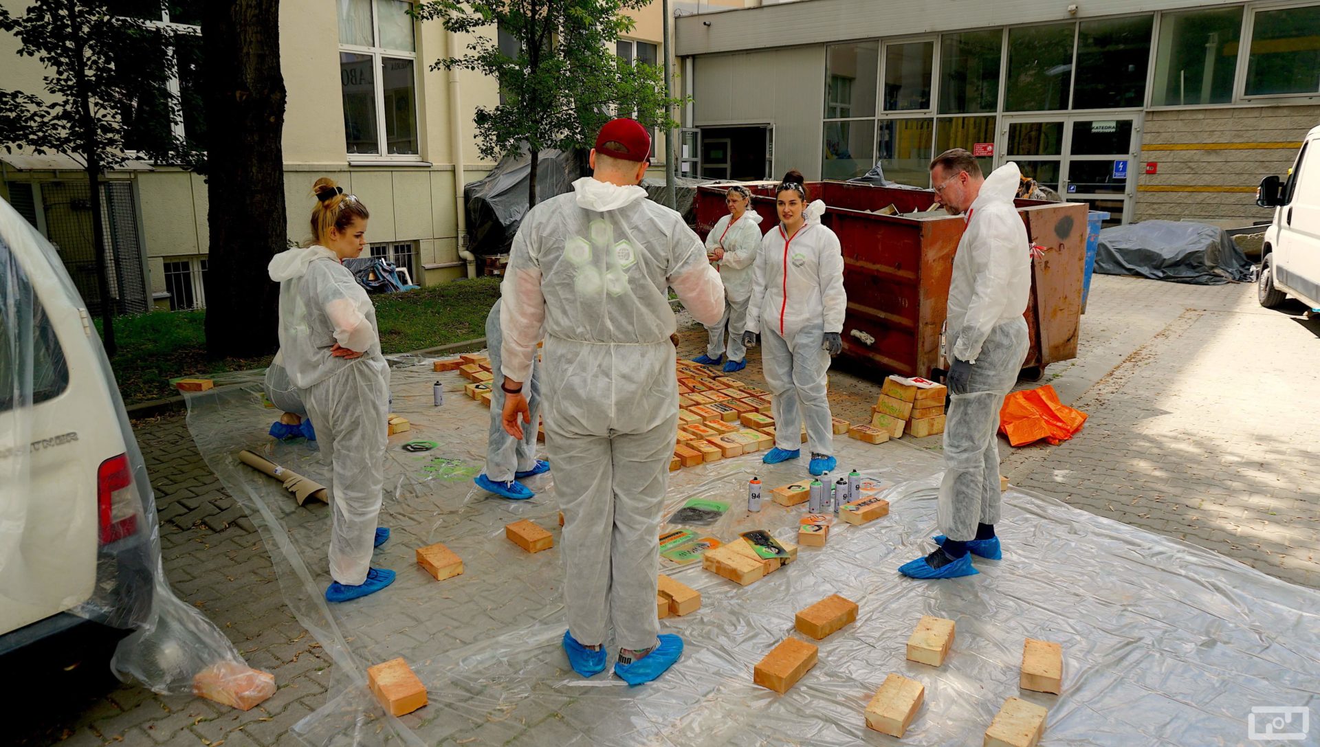Dziekan Wydziału prof. Marek Cała oraz trzy kobiety i jeden mężczyzna stoją ubrani w stroje malarskie i oglądają pomalowane cegły. Na rozłożonej folii malarskiej leżą pomalowane cegły oraz spraye.