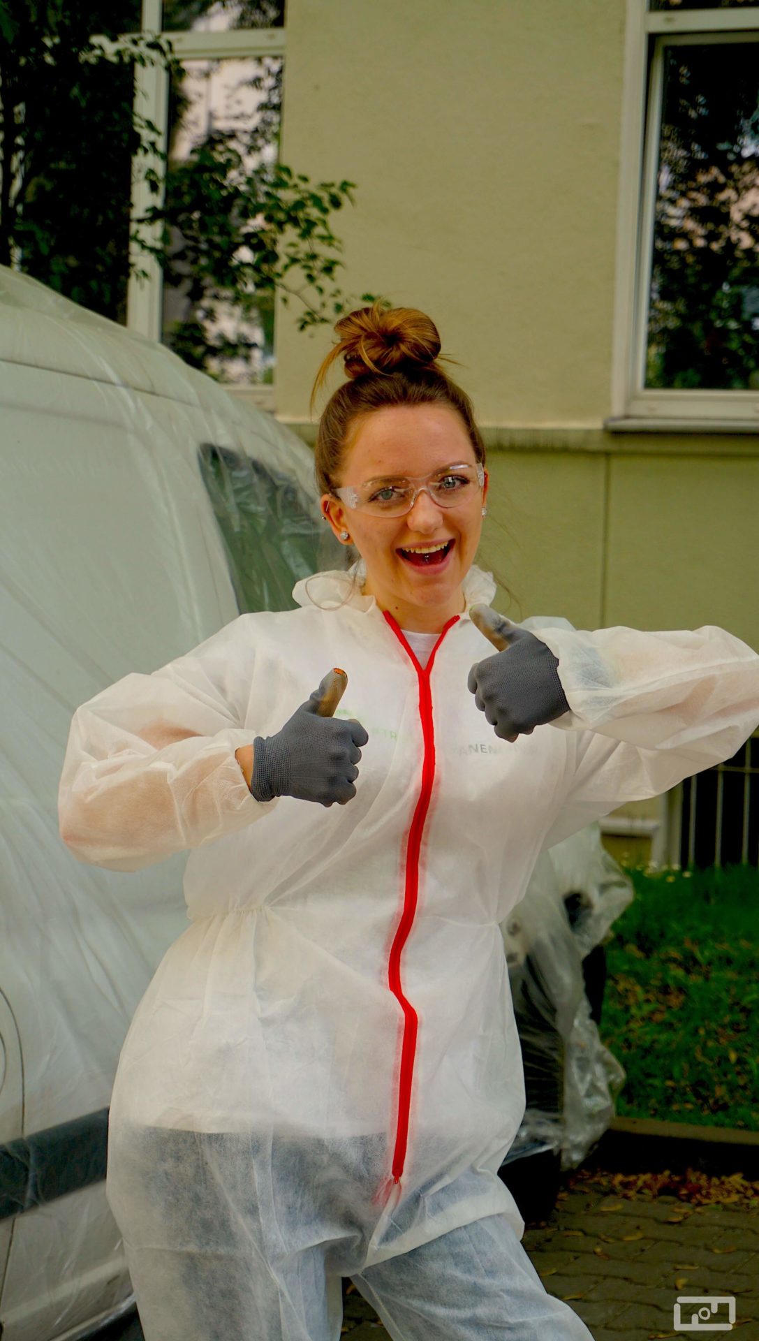 Uśmiechnięta studentka ubrana w strój malarski oraz gogle ochronne trzyma podniesione oba kciuki w górkę. Za nią budynek oraz okryty folią malarską samochód.