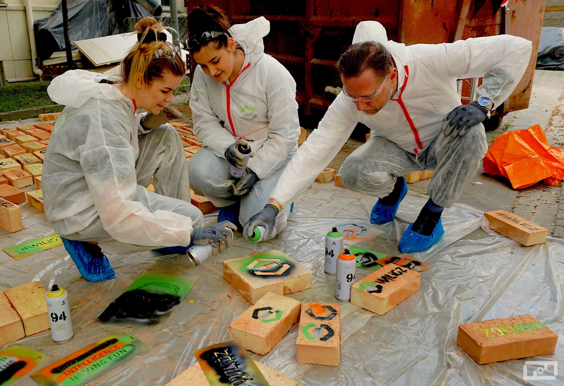 Dziekan Wydziału WILiGZ prof. Marek Cała wraz z dwoma studentkami ubrani w folie malarskie malują cegły przy użyciu sprayów i szablonów. Za nimi na foli leżą pomalowane cegły.