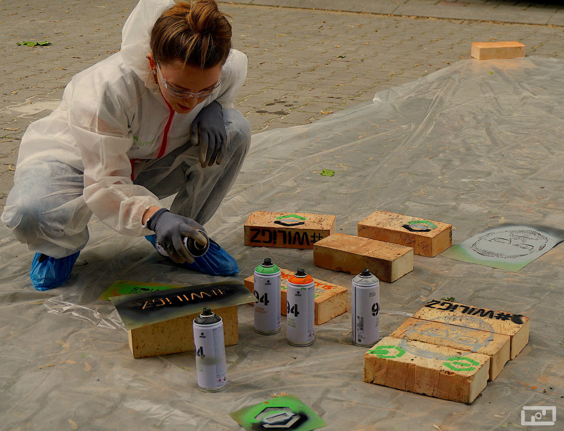 Uśmiechnięta młoda kobieta ubrana w strój malarski i gogle ochronne przy użyciu sprayu oraz szablonu maluje cegły. Przed nią na foli malarskiej leżą już pomalowane cegły.