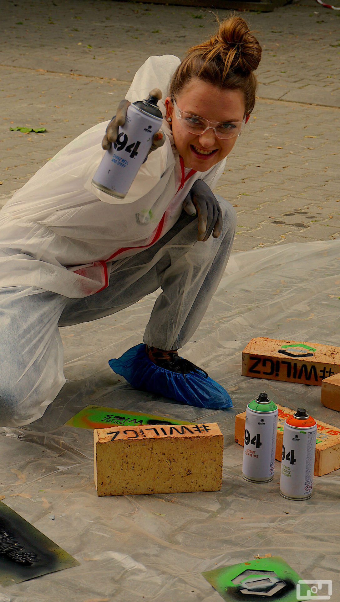 Uśmiechnięta młoda kobieta ubrana w strój malarski i gogle ochronne wyciąga ręke ze sprayem w kierunku aparatu. Przed nią na foli malarskiej leżą pomalowane cegły, spraye oraz szablony.