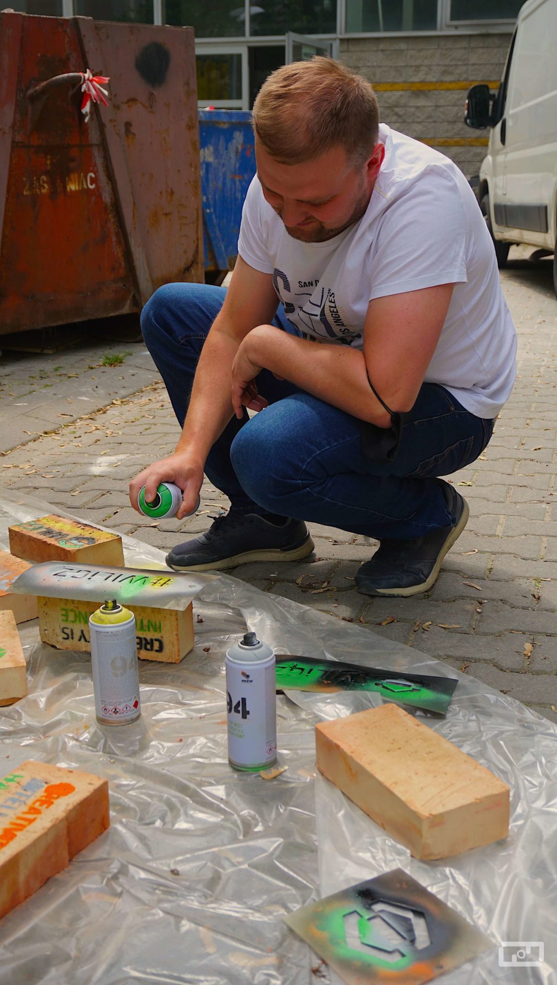 Mężczyzna maluje przy użyciu sprayu oraz szablonu leżące przed nimi na foli malarskiej cegły.
