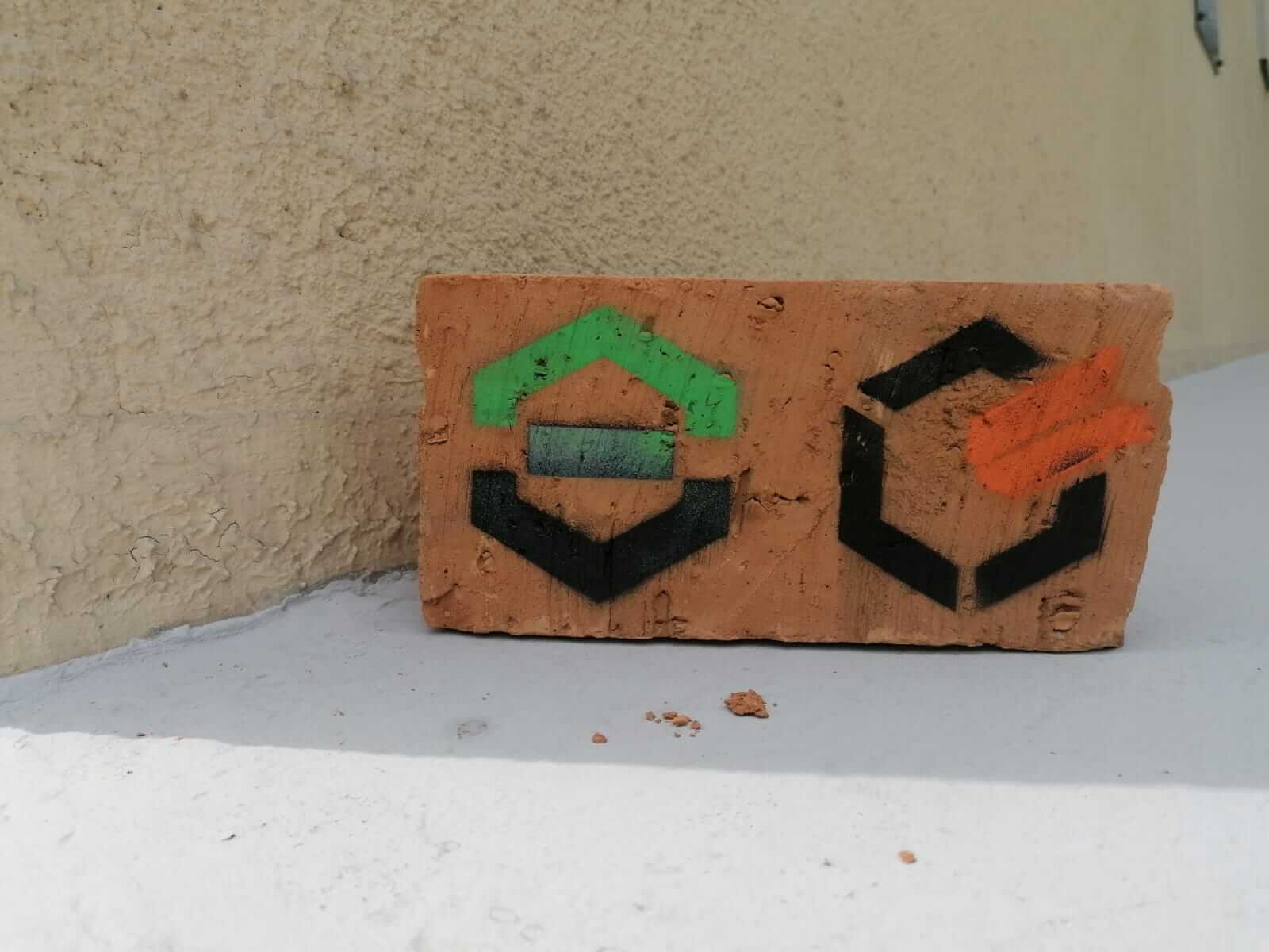 Pomarańćzowa cegła położona na szarym murku. Na lewej połówce cegły narysowane jest logo AGH na prawej połówce logo WRSS.