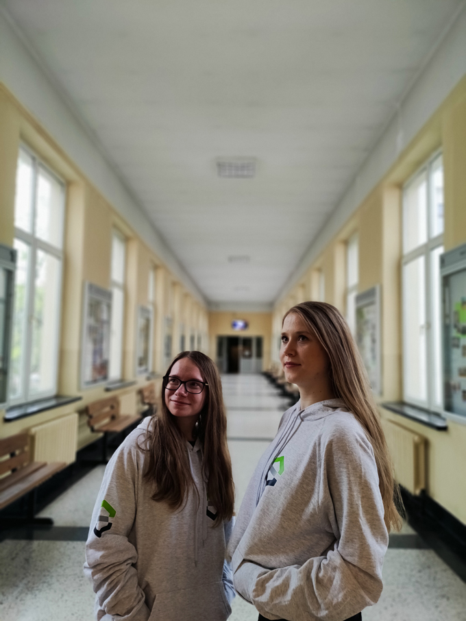 Dwie uśmiechnięte studentki stoją w bluzach wydziałowych na środku korytarza spoglądając w lewą stronę.