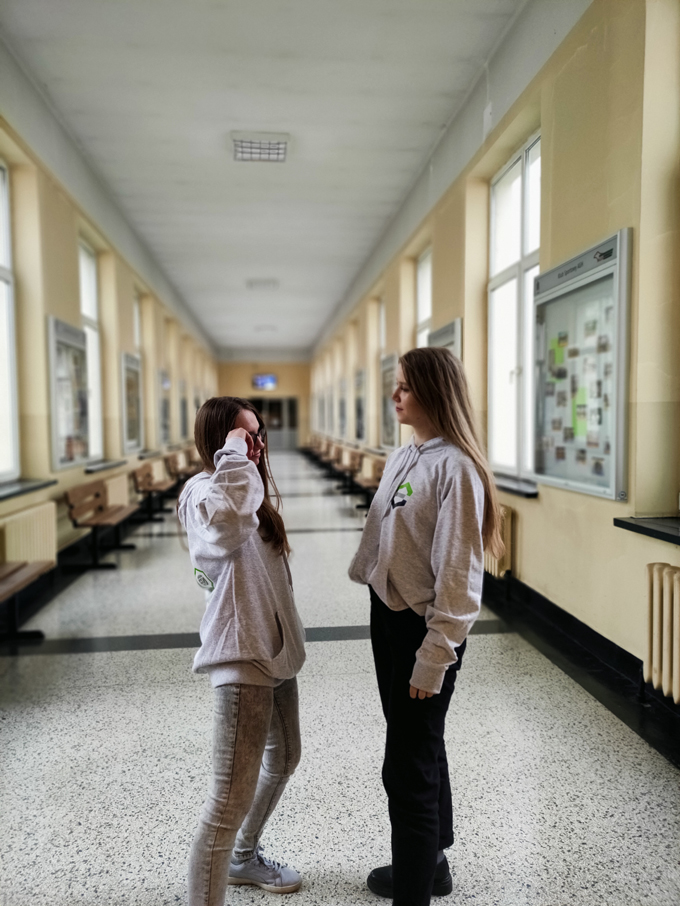Dwie uśmiechnięte studentki stoją w bluzach wydziałowych na środku korytarza. pięc symboli kierunków zamknięty w zielonych heksach.