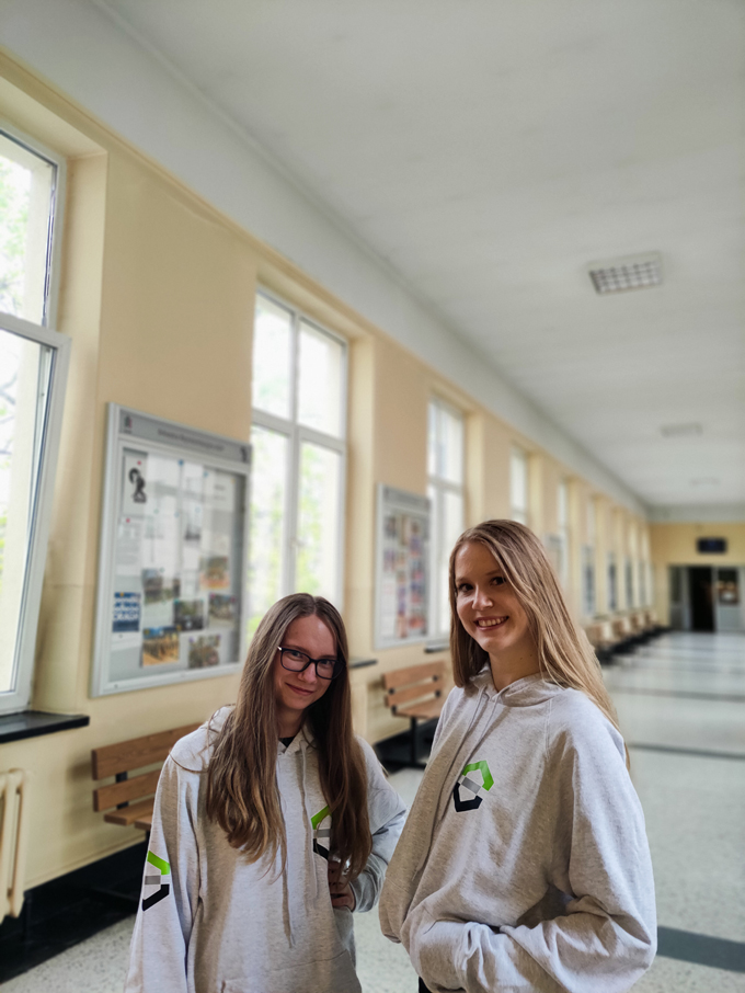 Dwie uśmiechnięte studentki stoją w bluzach wydziałowych na środku korytarza. pięc symboli kierunków zamknięty w zielonych heksach.