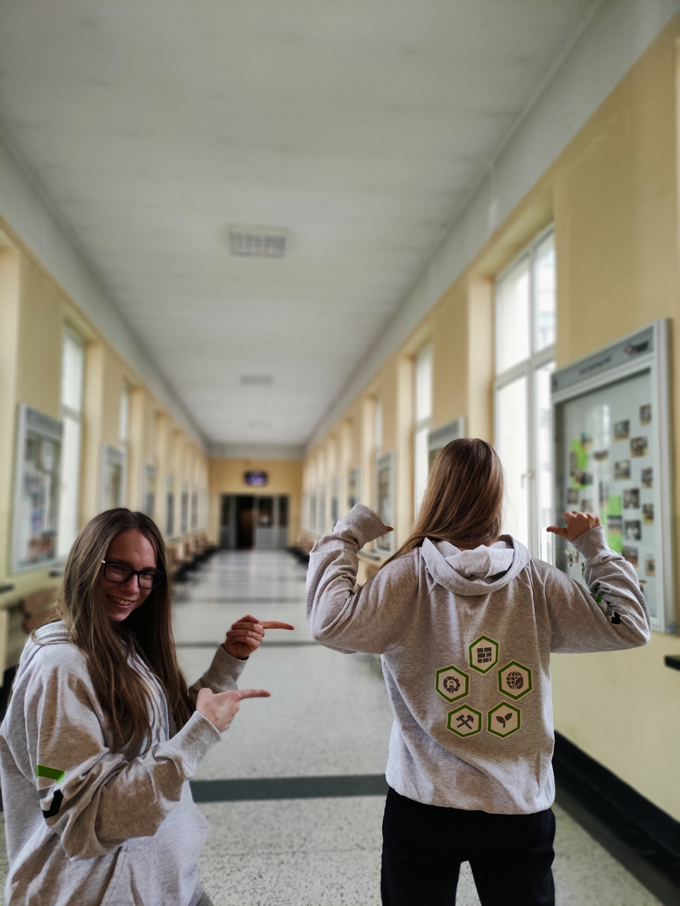 Dwie studentki w bluzach wydziaowych pozują stijąc w korytarzu. Studetka po lewej usmiecha się pokazując koleżankę Studentka po prawej obrócona jest plecami. Na plecach bluzy znajduje się pięc symboli kierunków.