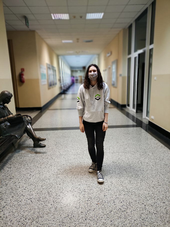 Studentka w szarej bluzie wydziałowej oraz długich czarnych włosach stoi uśmiechając się na korytarzu w budynku.
