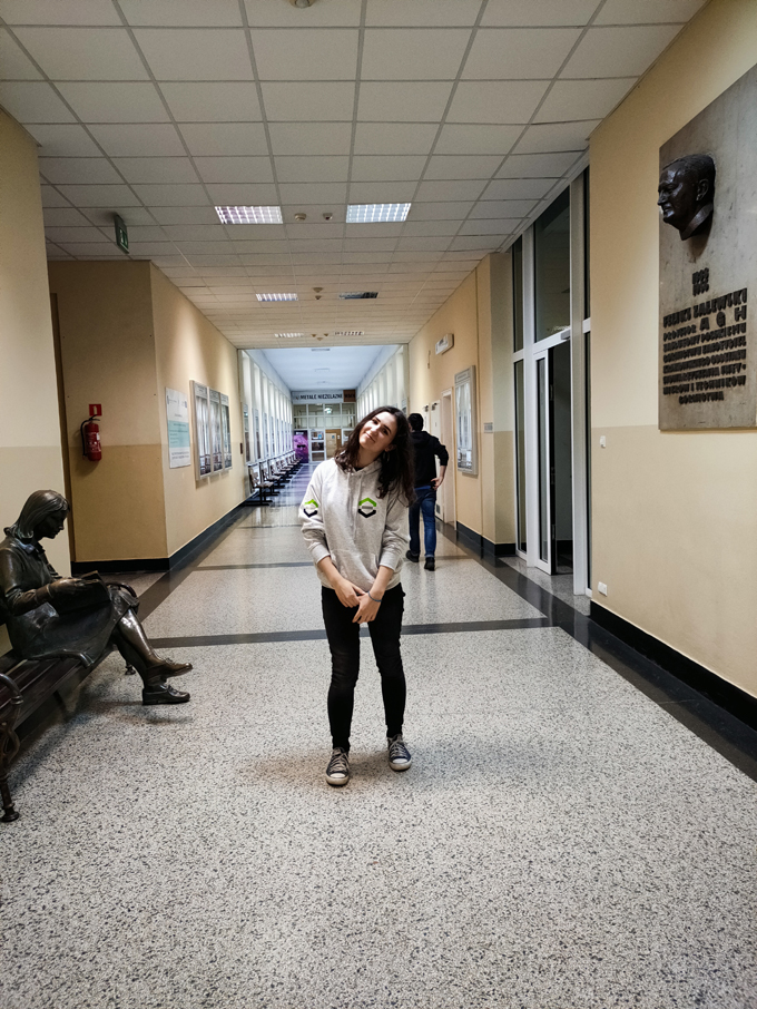 Studentka w szarej bluzie wydziałowej oraz długich czarnych włosach stoi uśmiechając się na korytarzu w budynku.