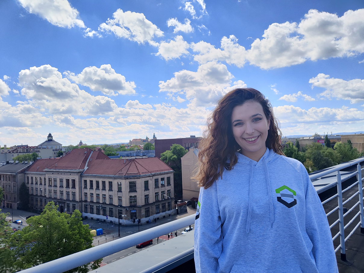 Uśmiechnięta studentka stoi w szarej bluzie na której jest logo wydziału przy balustradzie dachu. W tle widok na Stare Miasto Krakowa.