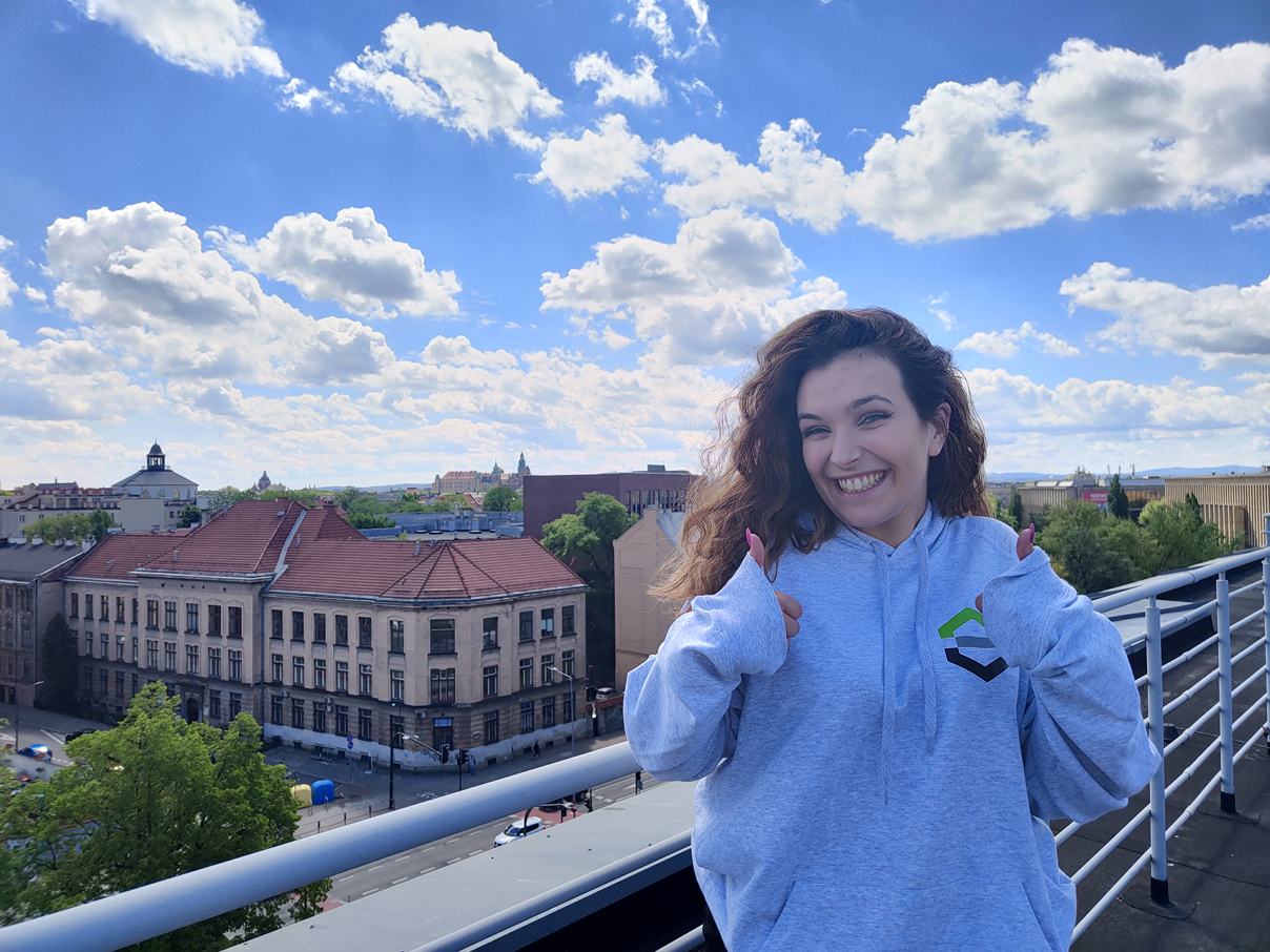 Uśmiechnięta studentka stoi w szarej bluzie na której jest logo wydziału przy balustradzie dachu trzymając podniesione w górę kciuki. W tle widok na Stare Miasto Krakowa.