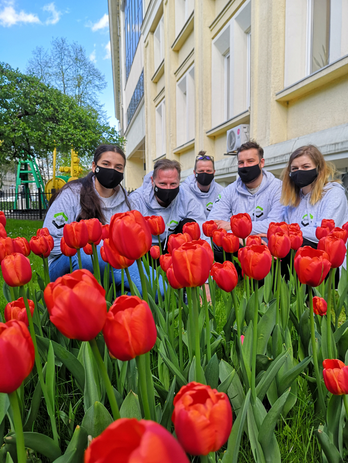 Dwie kobiety oraz trzech mężczyzn kucają przed rabatą z kwitnącymi czerwonymi tulipanami. Wszyscy ubrani sa w szare bluzy wydziałowe z czarnymi maseczkami na twarzy. Za ich plecami znajduje się budynek w kolorze piasku.