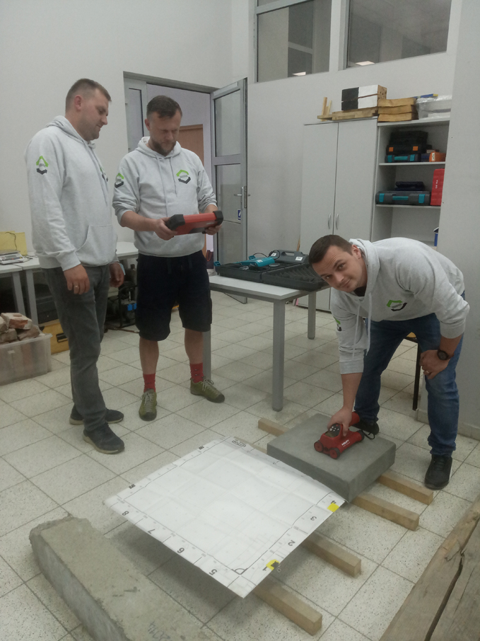 Trzeh pracowników wydziału ubranych w szare bluzy wydziałowe dokonują pomiaru betonowej płyty umieszczonej na podłodze laboratorum.