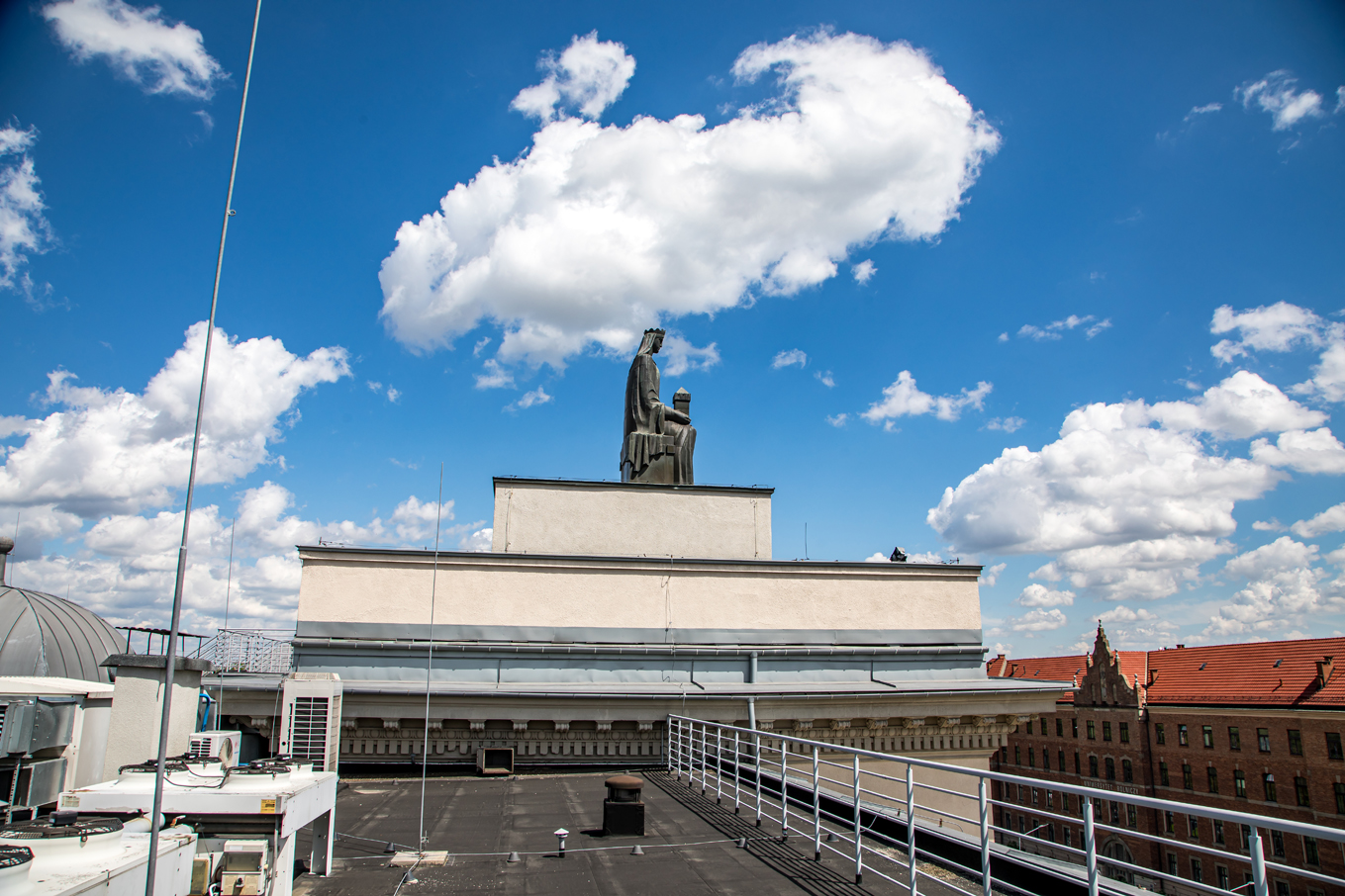 Pomnik świętej Barbary na tle błękitnego nieba. Poniżej pomnika czarny dach, balustrady, srebrne urządzenia klimatyzacyjne. Ujęcie z boku.