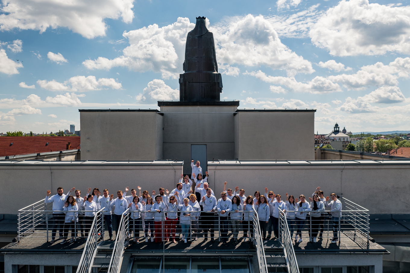 Pracownicy WILiGZ stoją na dachu budynku ubrani w jednolite szare bluzy wydziałowe i machają ręką na powitanie. W tle widać niebo oraz panorame Krakowa.