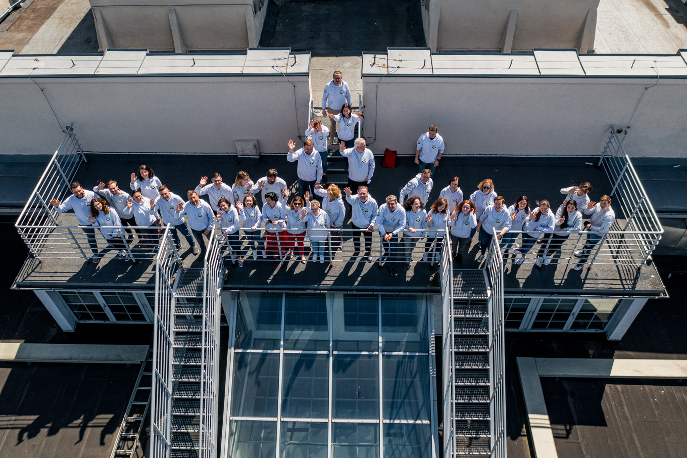 Pracownicy WILiGZ stoją na dachu budynku ubrani w jednolite szare bluzy wydziałowe i machają ręką na powitanie.