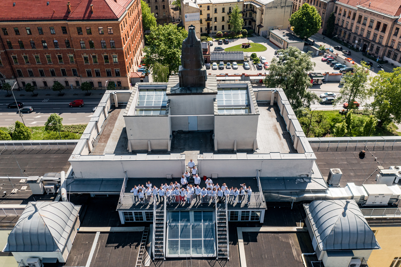Pracownicy WILiGZ stoją na dachu budynku ubrani w jednolite szare bluzy wydziałowe i machają ręką na powitanie. W tle widać niebo oraz panorame Krakowa.