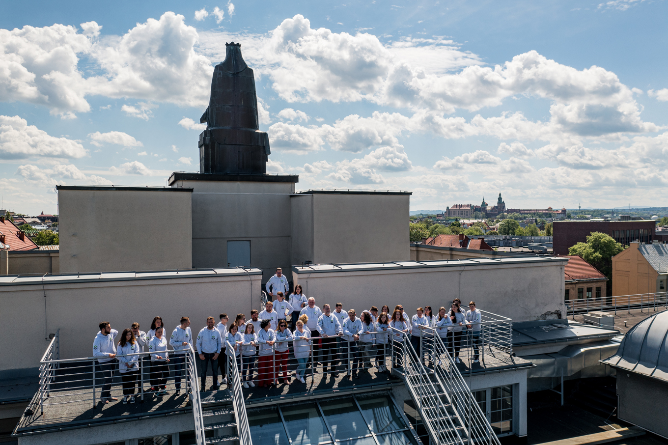 Pracownicy WILiGZ stoją na dachu budynku ubrani w jednolite szare bluzy wydziałowe. W tle widać niebo oraz panorame Krakowa.
