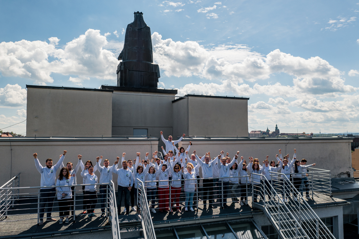 Pracownicy WILiGZ stoją na dachu budynku ubrani w jednolite szare bluzy wydziałowe z podniesionymi rękoma. W tle widać niebo oraz panorame Krakowa.