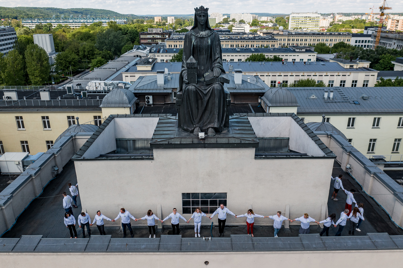 Pracownicy WILiGZ stoją wokół dachu budynku trzymając się za ręce . W tle widać czarny posąg Św. Barabry oraz dachy budynków AGH