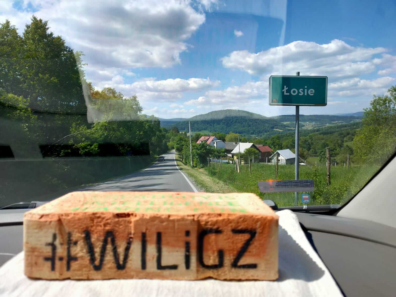 Cegła z czarnym hszatagiem i napisem WILiGZ leży na desce rozdzielczej samochodu. Przez szybe widać góry, domy, jezdnie oraz znak z nazwą miejcowości Łosie.