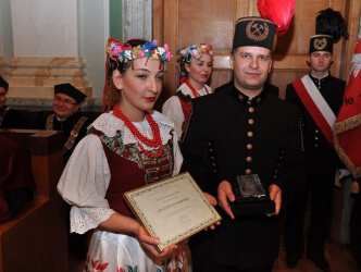Dwie kobiety ubrane tradycyjne strone krakowskie w towarzystwie 2 mężczyzn w mundurach górniczych. Jedna z kobiet niesie w rękach dymplom.