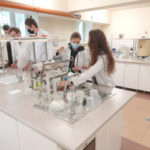 Studenci w białych chałatach ochronnych przy stołach na laboratirum.