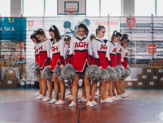Cheerleaderki ubrane w biało czerwone stroje z napisem AGH przygotowują się na układu na środku boiska. W rękach trzymają srebrne pompony.