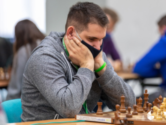 Mężczyzna ubrany w szarą bluzę i zieloną opaskę na rece zastanawia się nad szachownicą nad swoim kolejnym ruchem.