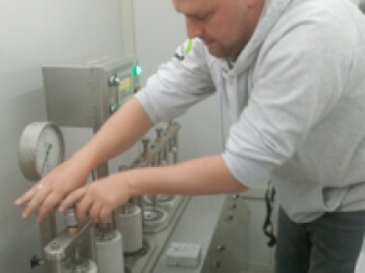 Mężczyzna w szarej bluzie wydziałowej wkłada próbki do stalowego urządzenia.