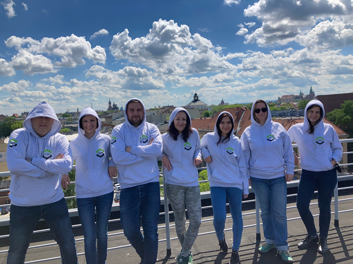 Pięć uśmiechniętych kobiet i dwóch mężczyzn stoją oparci o balustradę balkonową. Wszyscy ubrani są w szare bluzy wydziałowe z ubranymi kapturami na głowę. Za nimi w tle panorama starego miasta Krakowa oraz Zamek na Wawelu.