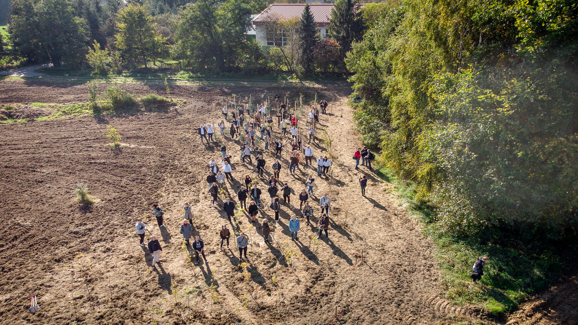 Studentki, studenci, Pracownicy oraz dzieci z pobliskiej szkoły z łopatami i sadzonkami w ręce na działce gdzie będzie prowadzona akcja. Zdjęcie z góry.
