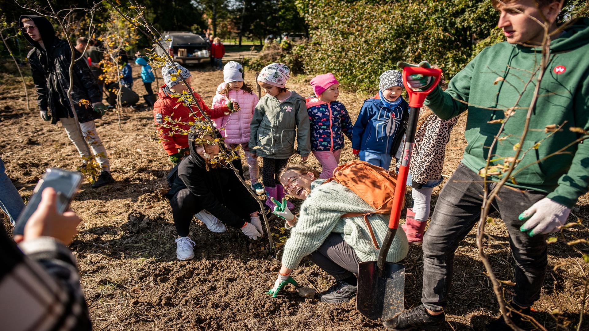 ką kolorowo ubranych dzieci przy zasadzonym przez nich drzewku. W tle studenci podczas sadzenia innych sadzonek.