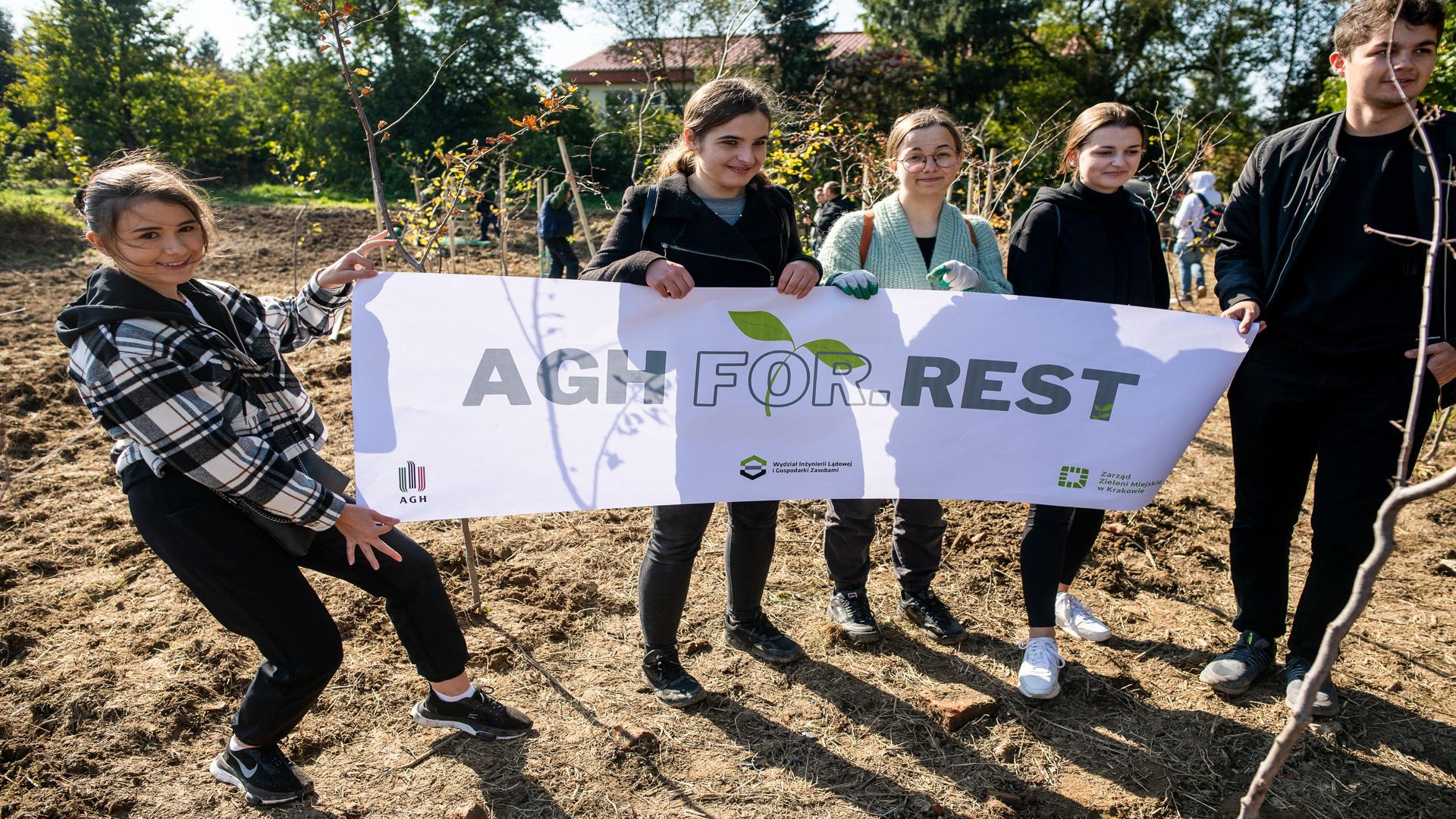 Cztery umiechnięte studentki oraz jeden student stoją obok siebie na polanie trzymając w ręce biały baner z szarym napisem AGH For.Rest. W tle zarys szkoły pomiędzy drzewami.