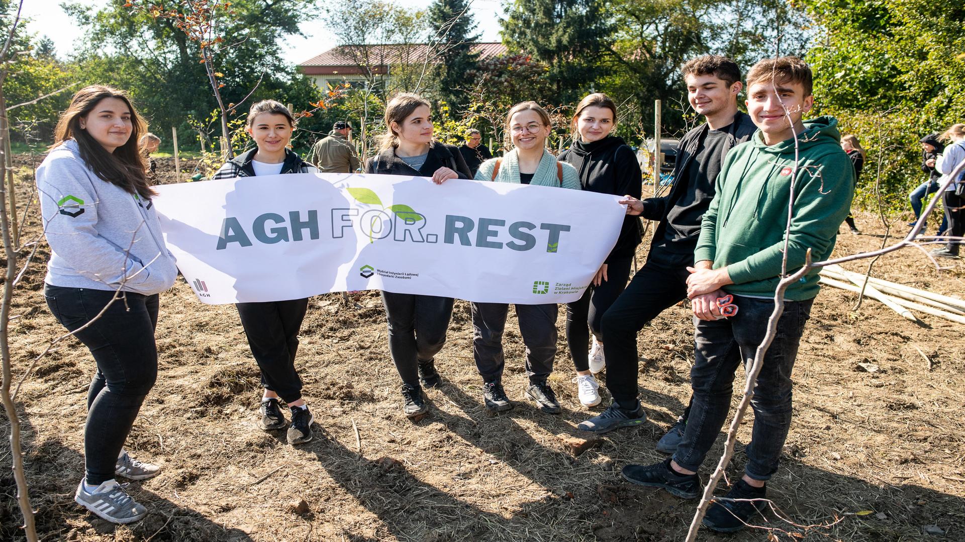 Cztery studentki oraz dwaj studenci stoją na polanie z białym banerem na którym znajduje się szary napis AGH For.Rest. Za nimi znajduje się budynek szkoły przysłonięty drzewami.