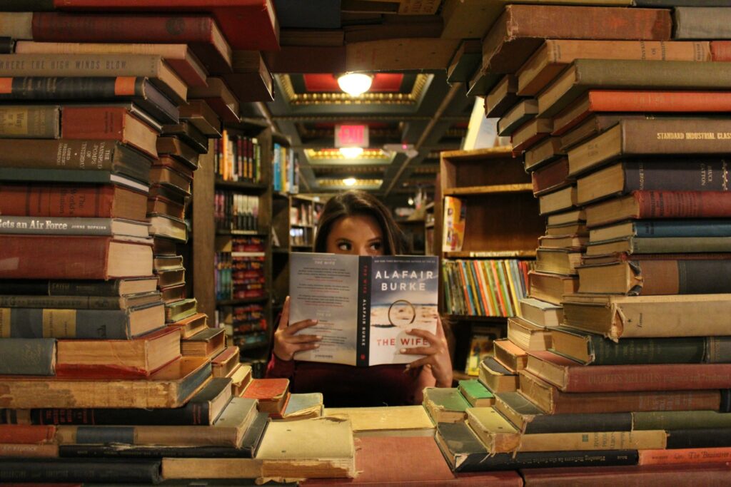 Kobieta czytająca książkę w Bibliotece. Stosy książek leżące na biurku po jej prawej i lewej stronie tworzą w środku okrąg przez który widziana jest kobieta. Za nią regały zastawione książkami.