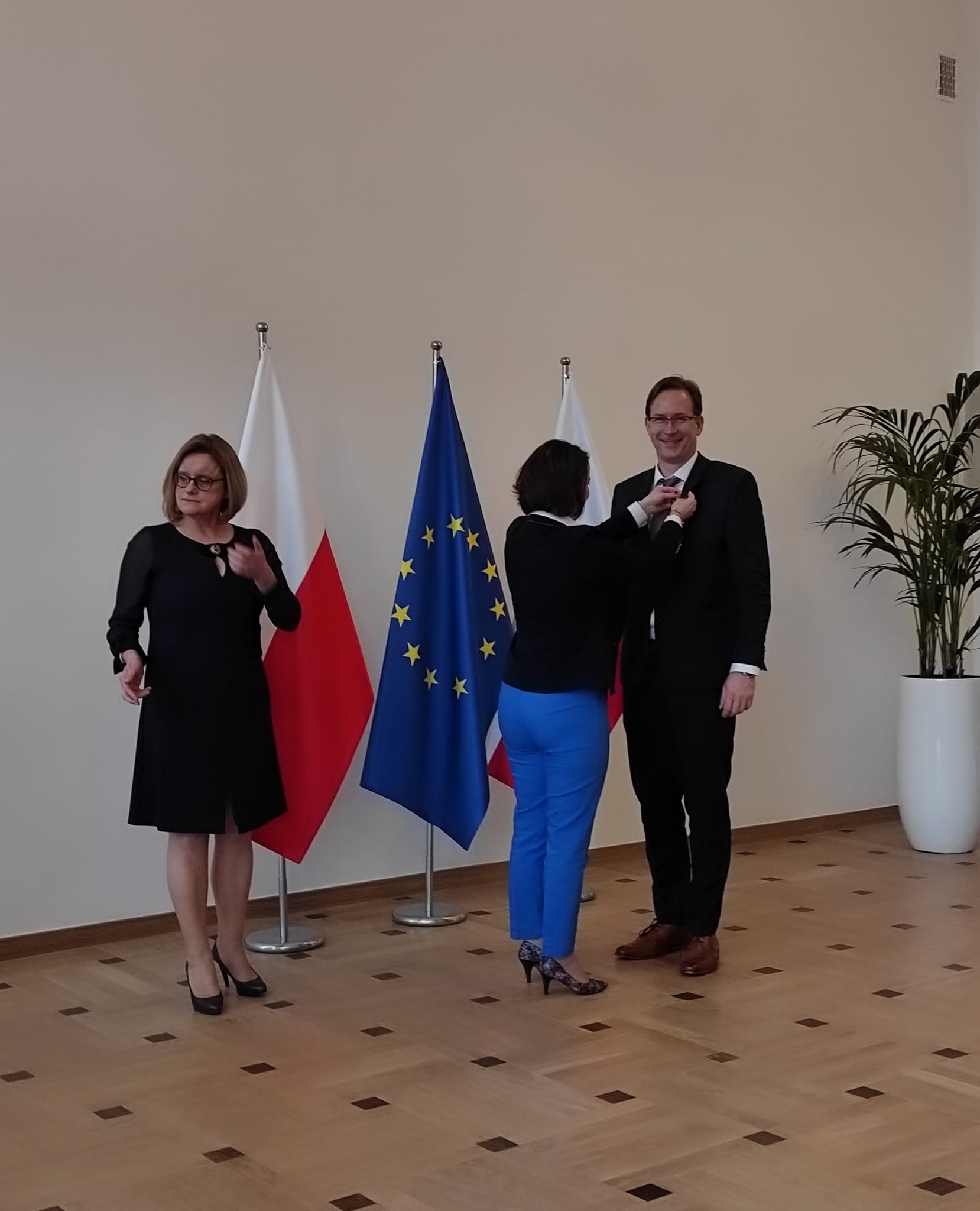 Ubrany na elegancko mężczyzana, stoi na baczność w dużej sali i odbiera nagrodę wręczaną przez kobietę. Za nim znajduje się dwie flagii Polski oraz jedna flaga Uni Europejskiej.