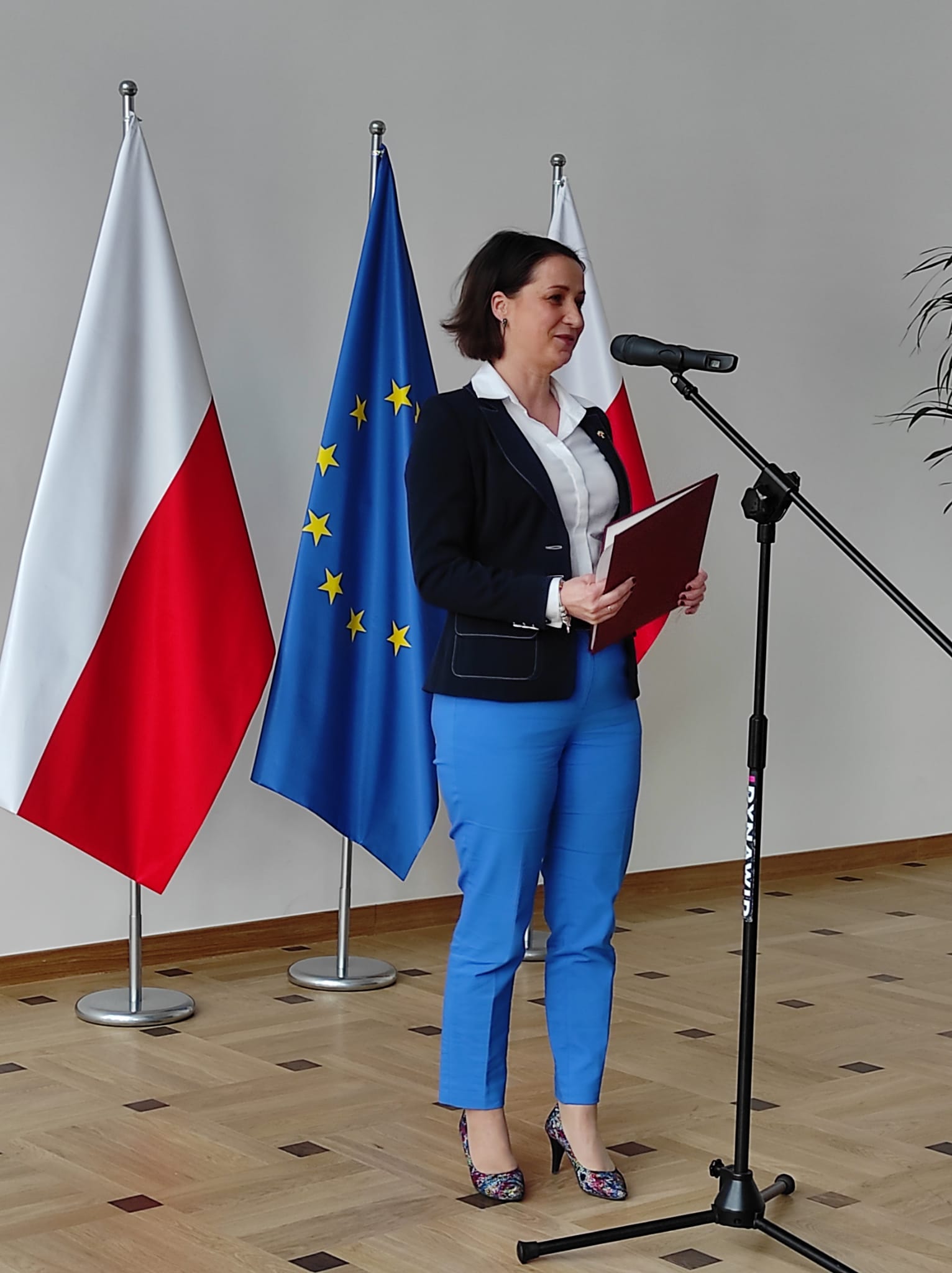 Elegancko ubrana kobieta stoi stoi przed mikrofonem, trzymając w ręku brązową kopertówkę. Za nią znajudją się dwie biało-czerwone flagii pomiędzy którymi znajuje się flaga flaga Unii Europejskiej.