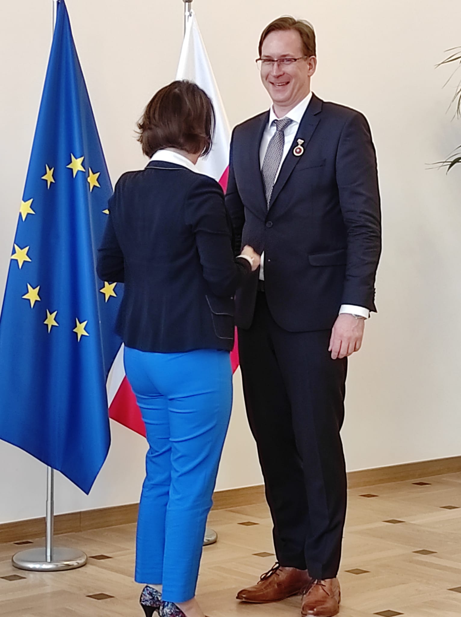 Uśmiechnięty mężczyzna ubrany w garnitur odbiera gratulacje od elegancko ubranej kobiety. Mężczyzna i kobieta stoją na tle flagi polski oraz Unii Europejskiej.