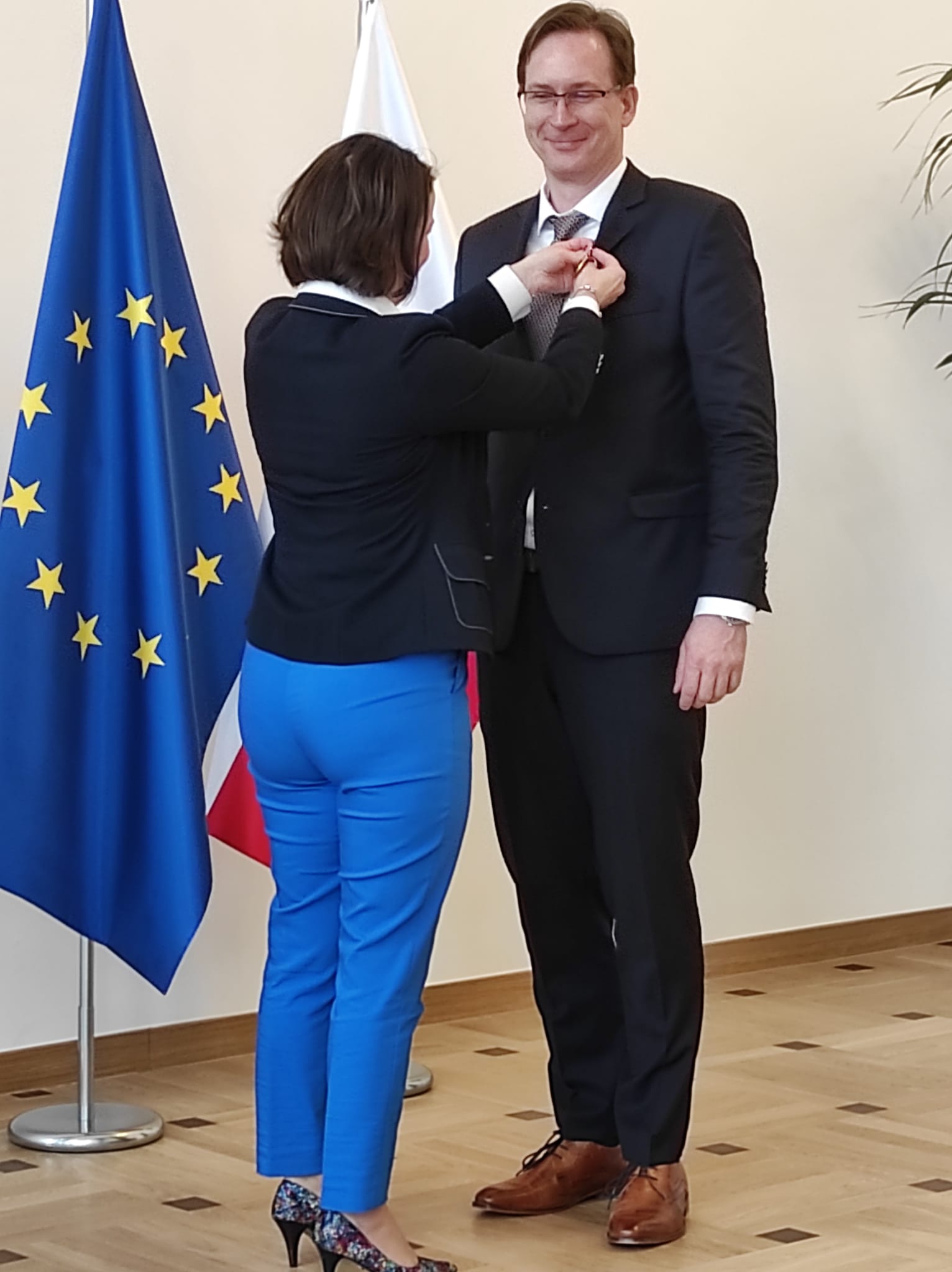 Uśmiechnięty mężczyzna ubrany w garnitur odbiera gratulacje od elegancko ubranej kobiety. Mężczyzna i kobieta stoją na tle flagi polski oraz Unii Europejskiej.