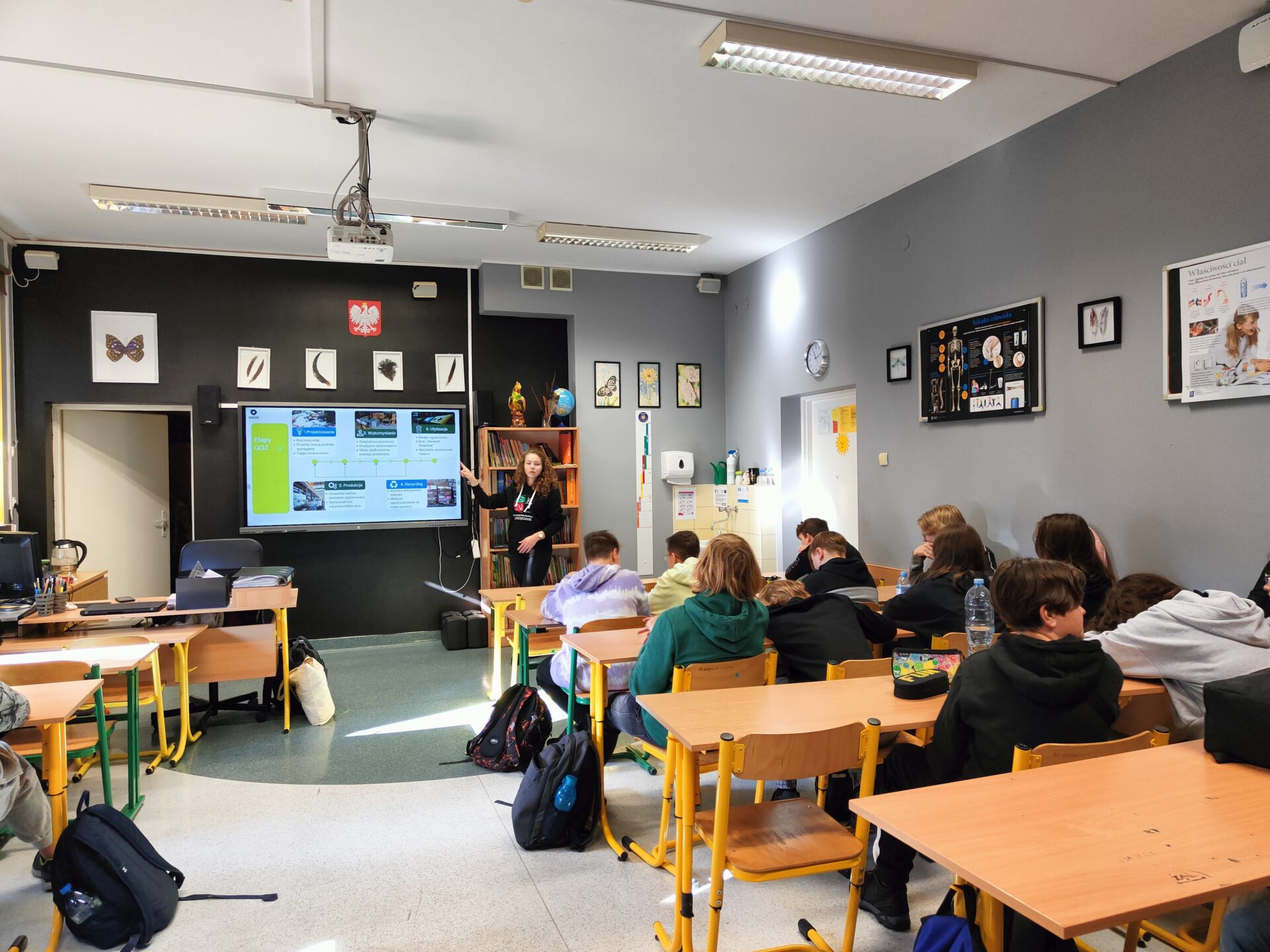 Uczniowie w ławkach w klasie. Z przodu studentka wskazująca na wyświetlony slajd prezentacji na ekranie.