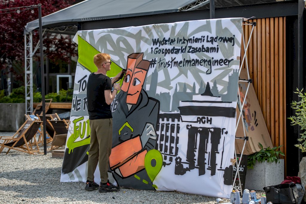 Młody mężczyzna sprayem maluje graffiti na rozwieszonym dużym płótnie